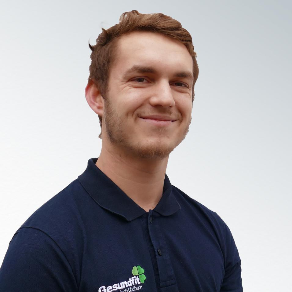 Junger Mann mit kurzem Haar, lächelnd, trägt ein dunkles Poloshirt mit dem Logo "Gesundfit". Heller Hintergrund.
