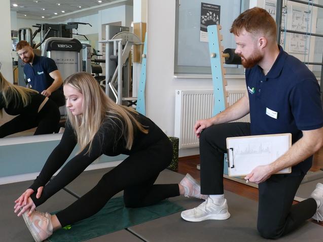 Frau dehnt sich auf einer Trainingsmatte, w&auml;hrend ein Trainer mit Notizen daneben kniet und sie anleitet.