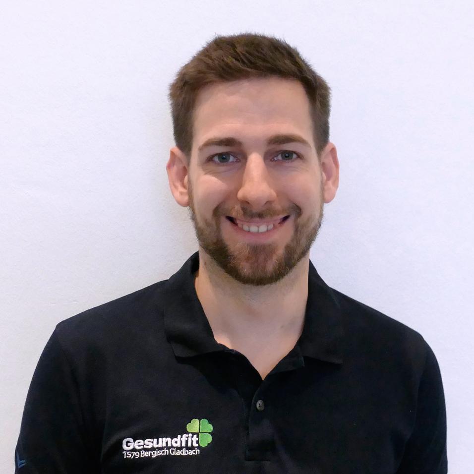 Porträt eines lächelnden Mannes mit kurzer Frisur, schwarzem Polo-Shirt mit Logo, vor einem hellen Hintergrund.