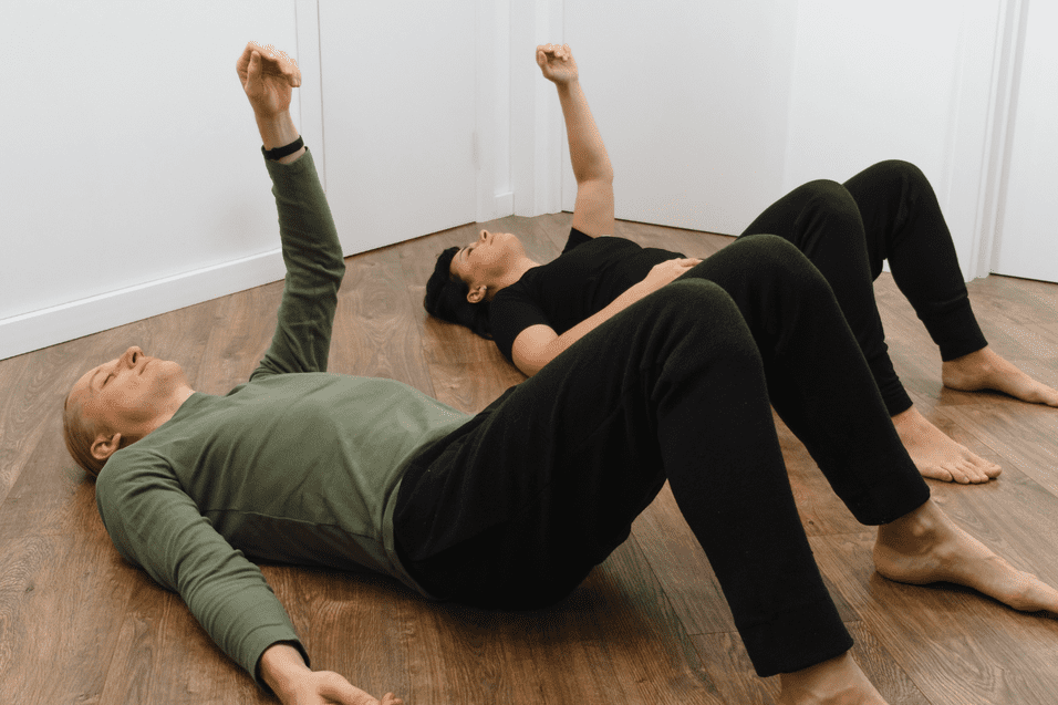 Zwei Personen liegen auf dem Boden, Arme erhoben, in einem hellen Raum mit Holzfu&szlig;boden.