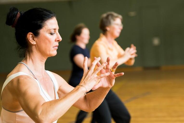 Drei Frauen in sportlicher Kleidung &uuml;ben Bewegungen in einer Turnhalle mit Holzfu&szlig;boden.