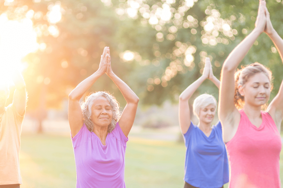 Vier Personen praktizieren Yoga im Freien, mit erhobenen H&auml;nden vor einer gr&uuml;nen Kulisse bei Sonnenaufgang.