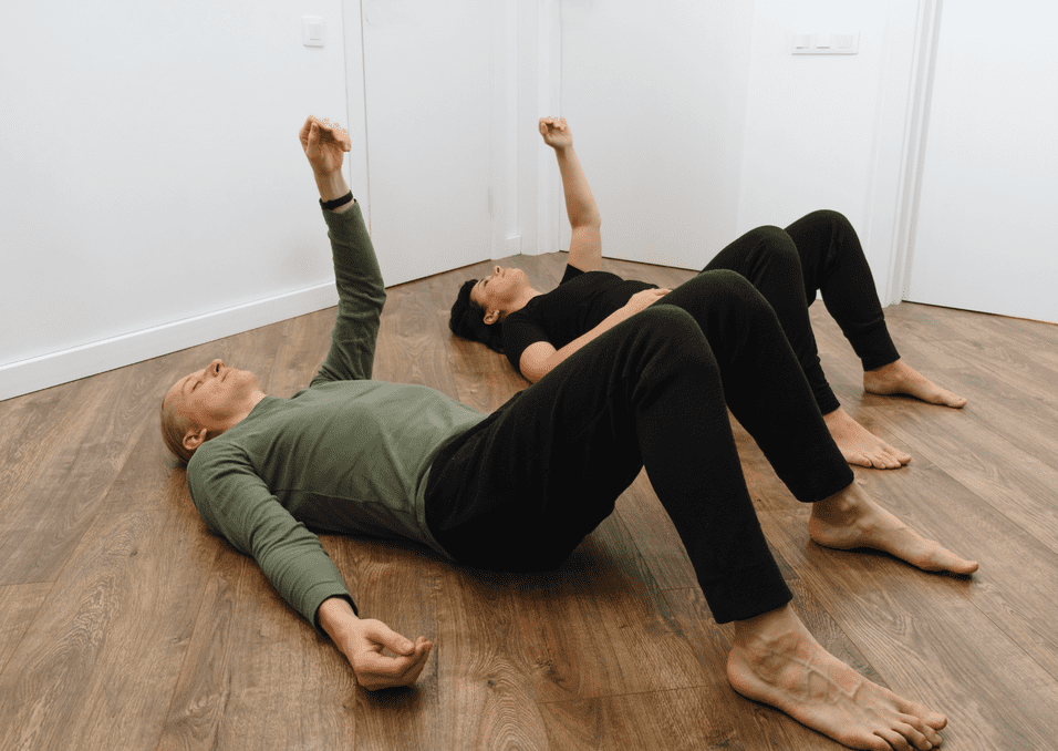 Zwei Personen liegen auf dem Boden, Arme erhoben, in einem hellen Raum mit Holzfu&szlig;boden.