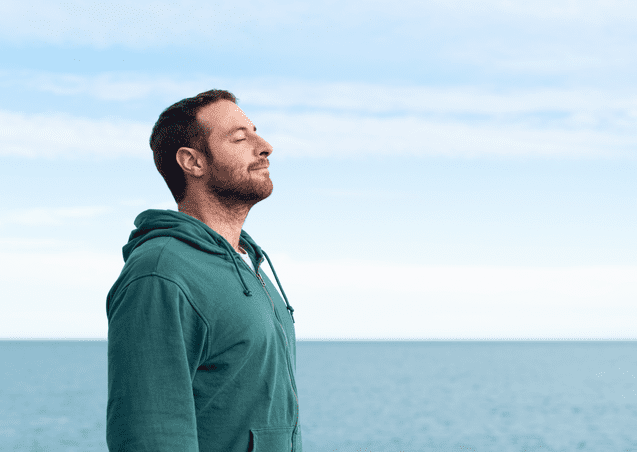 Mann mit geschlossenen Augen steht am Wasser und genie&szlig;t die frische Luft, Hintergrund zeigt einen blauen Himmel.