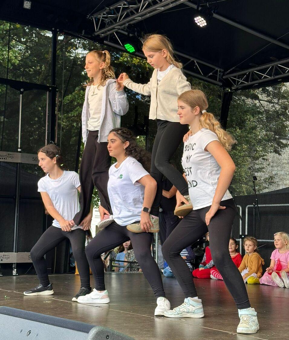 F&uuml;nf T&auml;nzerinnen vor einer B&uuml;hne in verschiedenen Positionen, w&auml;hrend Kinder im Hintergrund zuschauen.