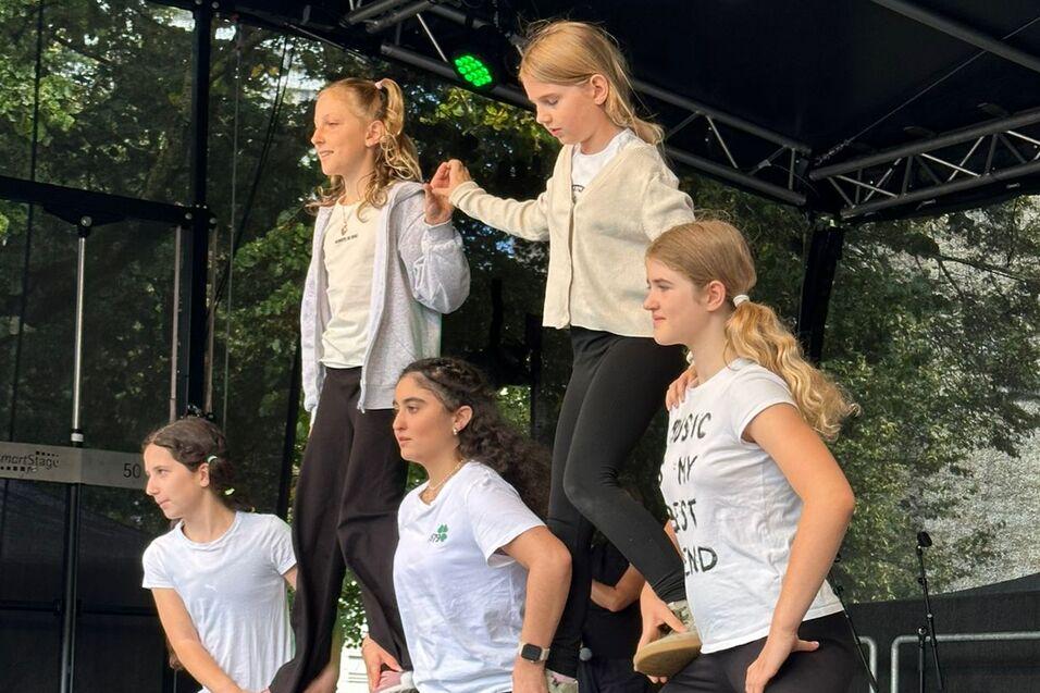 F&uuml;nf T&auml;nzerinnen vor einer B&uuml;hne in verschiedenen Positionen, w&auml;hrend Kinder im Hintergrund zuschauen.