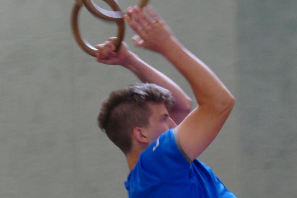 Junge trainiert an Turnringen, mit leicht angehobenem K&ouml;rper und einem sportlichen Outfit in Blau und Rot.