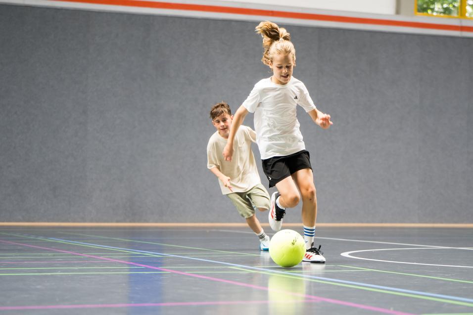 Zwei Kinder spielen in einer Halle Fu&szlig;ball, ein Kind rennt dem Ball hinterher, w&auml;hrend das andere zuschaut.