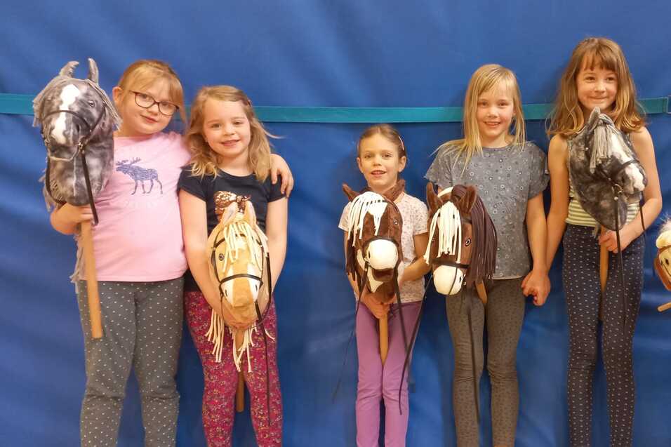 Sechs M&auml;dchen stehen in einer Reihe und halten Hobby-Pferde aus Stoff, vor einer blauen Wand im Hintergrund.