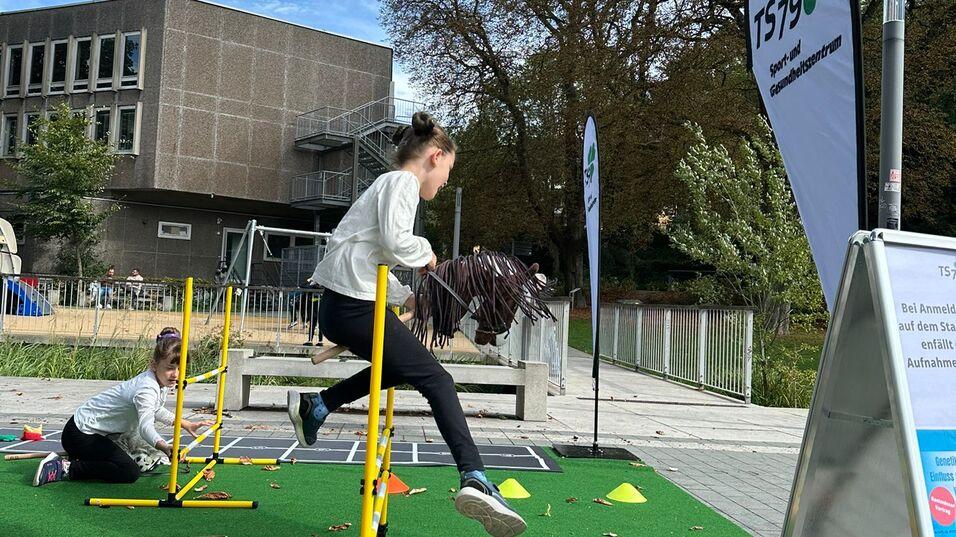 Zwei Kinder absolvieren einen Hindernisparcours im Freien, umgeben von gr&uuml;ner Mattenfl&auml;che und Trainingsmaterialien.