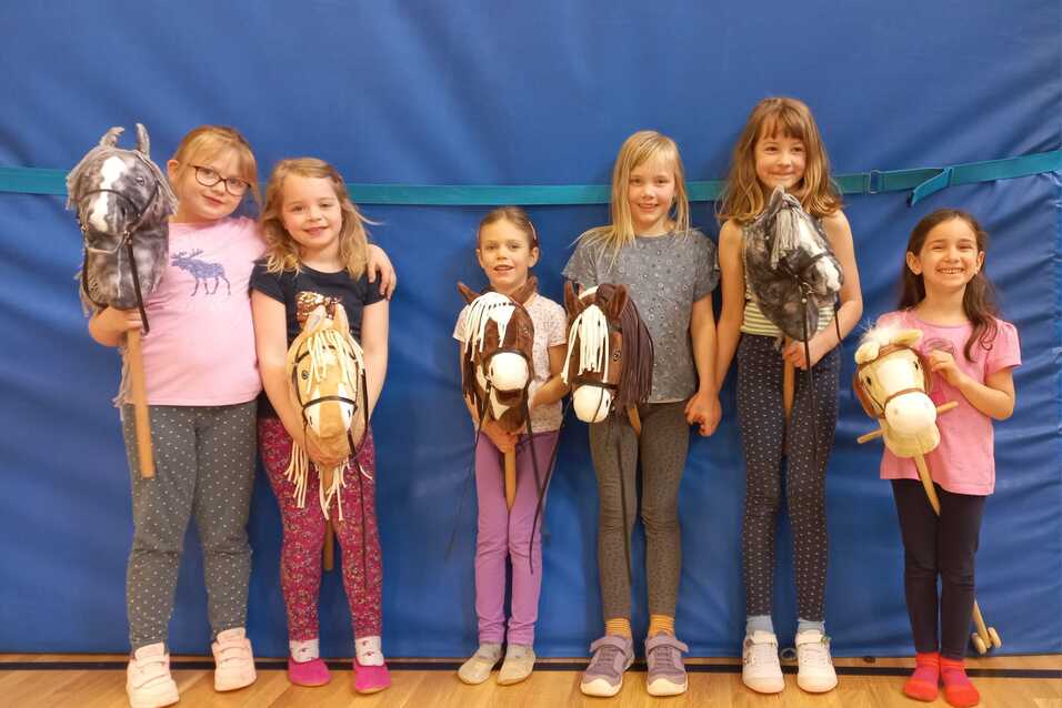 Sechs M&auml;dchen stehen in einer Reihe und halten Hobby-Pferde aus Stoff, vor einer blauen Wand im Hintergrund.