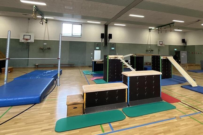 Blick in eine Sporthalle mit verschiedenen Turnger&auml;ten, Matten und einer Holzrampe auf dem hellen Parkettboden.