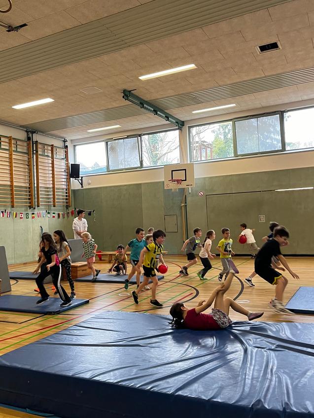 Kinder spielen in einer Turnhalle, hüpfen über Matten und benutzen Bälle zum Sport. Farbige Flaggen schmücken die Wände.