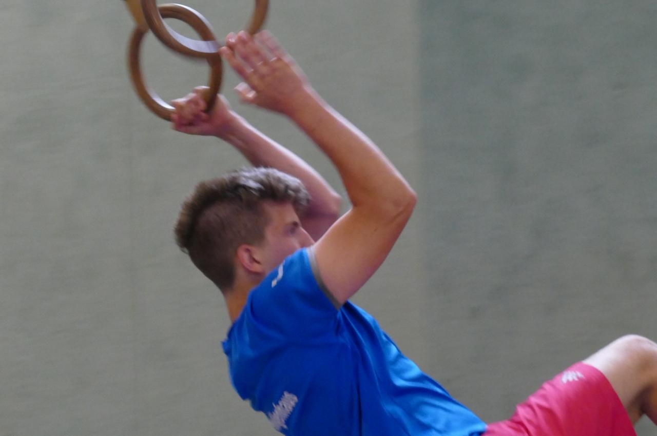 Junger Mann schwingt an Turnringen in blauer T-Shirt und roter Shorts an einer Wand mit gr&uuml;ner Farbe.
