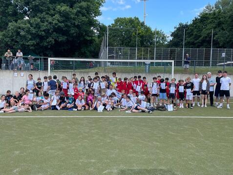 Gruppierung von Kindern und Jugendlichen in Sportkleidung auf einem Fußballfeld, umgeben von Zuschauern.