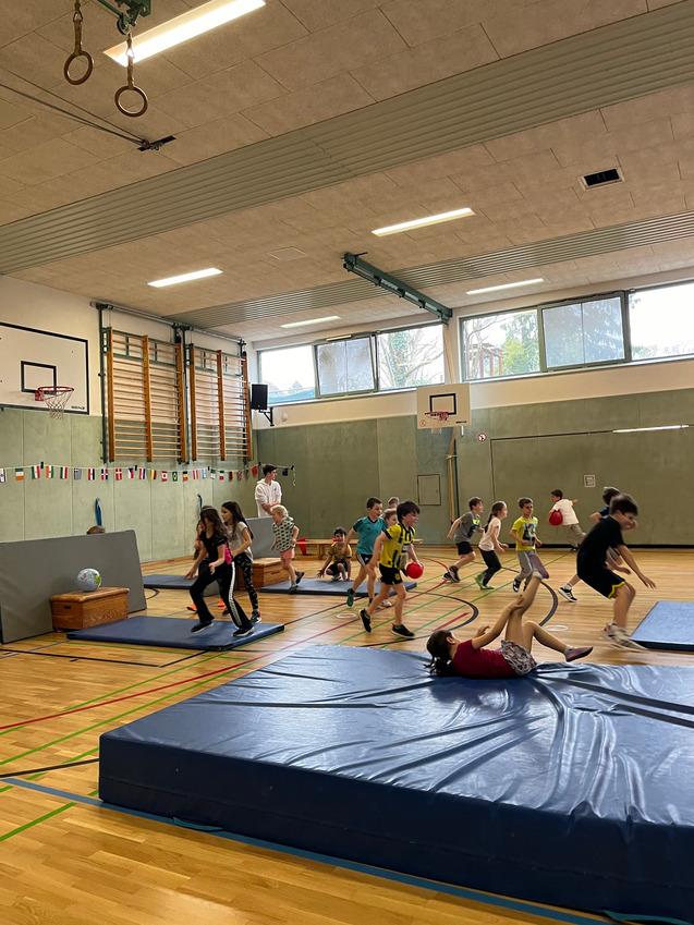 Kinder spielen in einer Turnhalle, h&uuml;pfen &uuml;ber Matten und benutzen B&auml;lle zum Sport. Farbige Flaggen schm&uuml;cken die W&auml;nde.