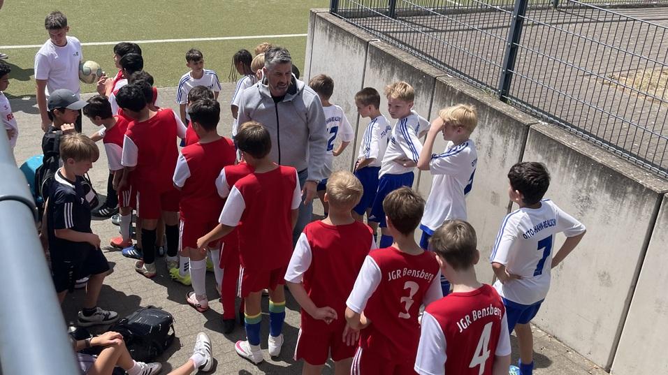 Gruppe von Kindern in rot-wei&szlig;en Trikots lauscht einem Trainer auf einem Fu&szlig;ballplatz mit Kunstrasen.