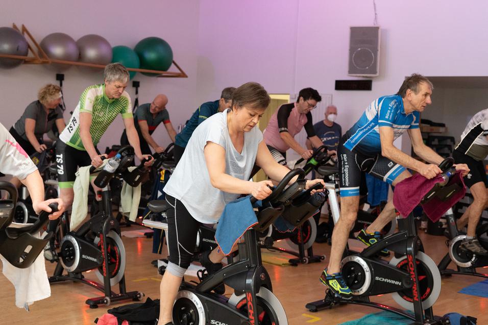 Gruppe von Personen beim Indoor-Cycling in einem Fitnessraum mit verschiedenen Fahrr&auml;dern und Fitnessger&auml;ten im Hintergrund.