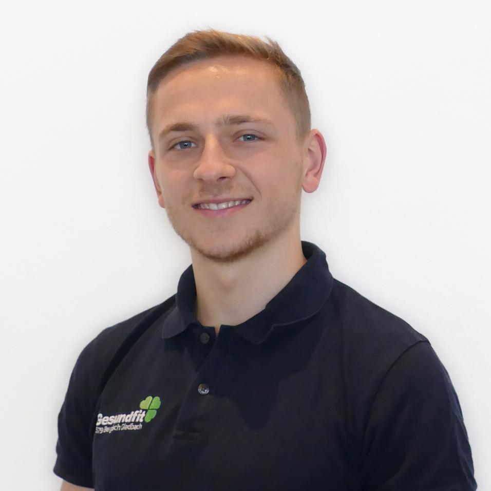 Portr&auml;t eines jungen Mannes mit kurzem, blondem Haar, der in einem schwarzen Poloshirt vor einer wei&szlig;en Wand steht.