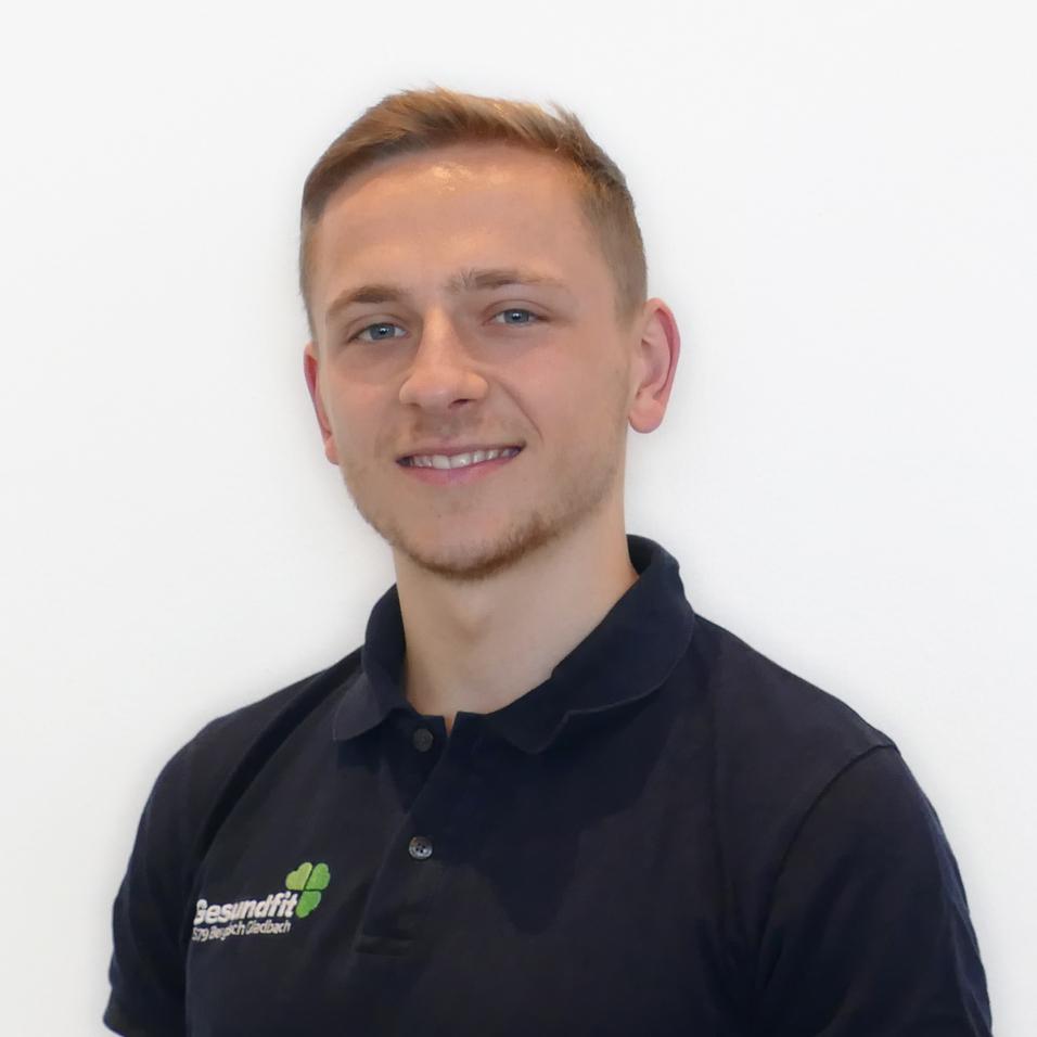 Porträt eines jungen Mannes mit kurzem, blondem Haar, der in einem schwarzen Poloshirt vor einer weißen Wand steht.