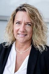 Porträt einer lächelnden Frau mit lockigem, blondem Haar, die ein schwarzes Blazer-Jacket und eine weiße Bluse trägt.
