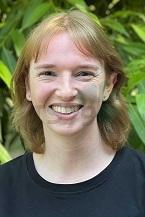 Lächelnde Person mit kurzen, blonden Haaren vor einer grünen, pflanzenreichen Hintergrundlandschaft.