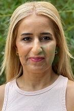 Porträt einer Frau mit langen blonden Haaren, trägt ein helles Oberteil, vor einem unscharfen grünen Hintergrund.