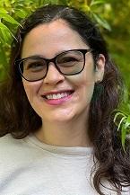 Porträt einer Frau mit Brille und lächelndem Gesicht, umgeben von grünen Pflanzen im Hintergrund.