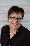 Porträt einer lächelnden Frau mit kurzen Haaren und Brille vor einem hellen Hintergrund. Sie trägt ein dunkles Oberteil.