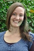 Frau mit langen, braunen Haaren lächelt vor einem Hintergrund aus grünen Blättern. Sie trägt ein blaues Oberteil.