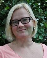 Porträt einer blonden Frau mit Brille, lächelnd vor einem Hintergrund aus grünen Blättern.