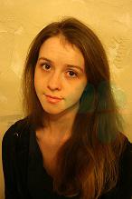 Portr&auml;t einer jungen Frau mit langen, braunen Haaren, neutralem Ausdruck vor einer hellen Wand.