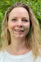 Portr&auml;t einer Person mit langen blonden Haaren vor einer gr&uuml;nen Pflanzenkulisse, l&auml;chelnd in die Kamera blickend.