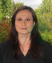 Portr&auml;t einer Frau mit langen, dunklen Haaren, vor einer gr&uuml;nen, bewaldeten Kulisse im Hintergrund.