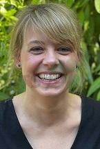 L&auml;chelnde Frau mit blonden Haaren vor gr&uuml;ner Pflanzenkulisse. Sie tr&auml;gt ein schwarzes Oberteil.