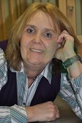 Frau mit blonden Haaren, im Freizeitlook, l&auml;chelt entspannt, w&auml;hrend sie auf einem Tisch liegt und ihr Kopf auf die Hand st&uuml;tzt.