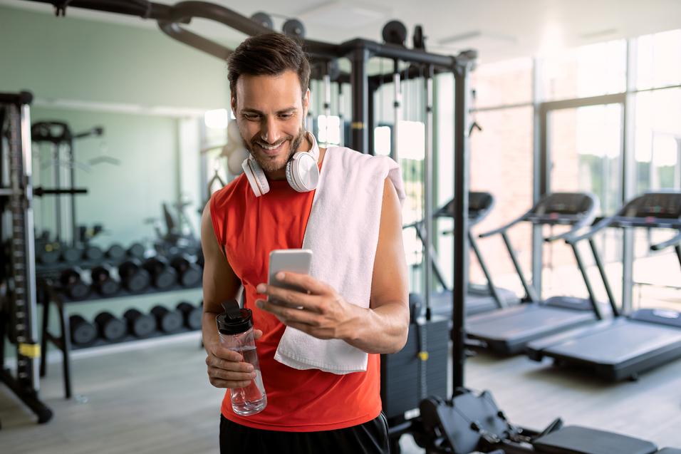 Mann in rotem Sportshirt mit Kopfh&ouml;rern und Handtuch, schaut l&auml;chelnd auf sein Smartphone im Fitnessstudio.