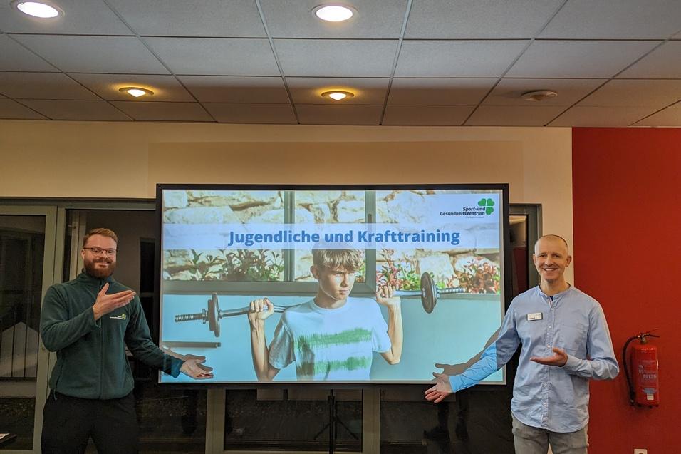 Zwei Männer präsentieren ein Bildschirmthema „Jugendliche und Krafttraining“ in einem hellen Raum.