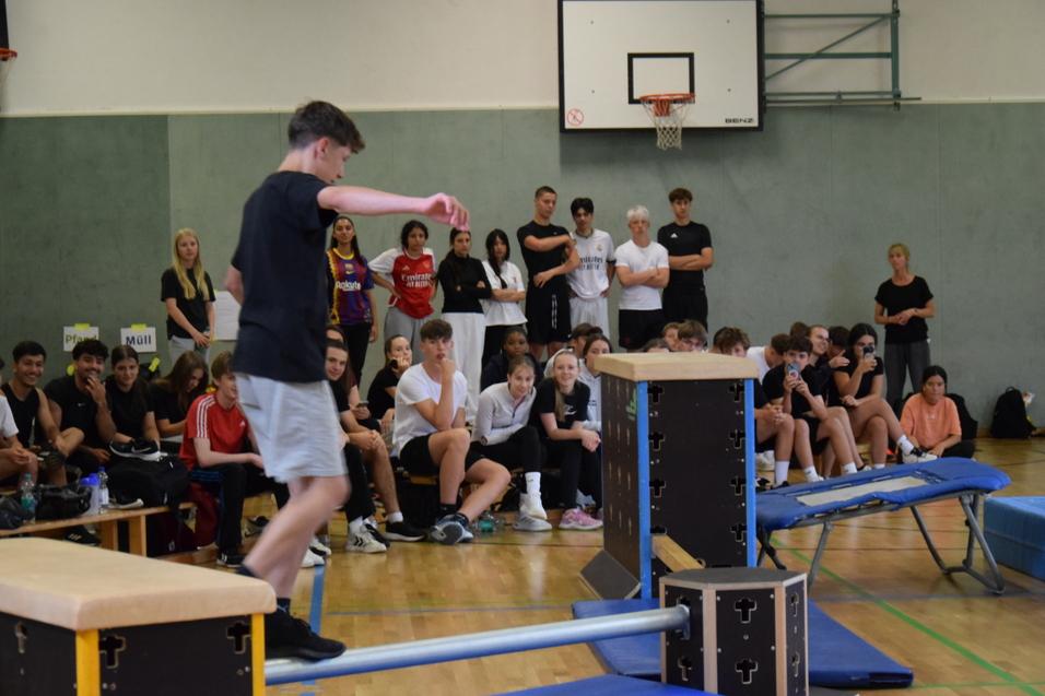 Ein Turner balanciert auf einem niedrigen Balken, während eine Gruppe Zuschauer zuschaut. Sporthalle im Hintergrund.