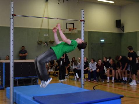 Ein Athlet in grüner T-Shirt springt über einen Hochsprungbalken in einer Sporthalle, Zuschauer beobachten.