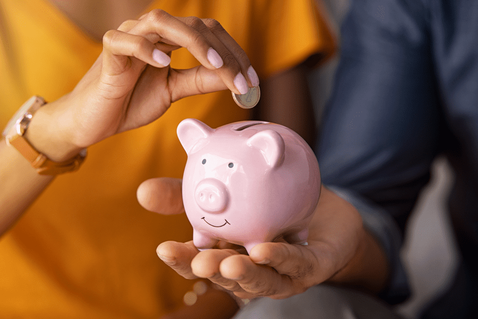 Nahaufnahme einer Hand, die eine M&uuml;nze in ein rosa Sparschwein steckt, w&auml;hrend eine andere Hand das Sparschwein h&auml;lt.