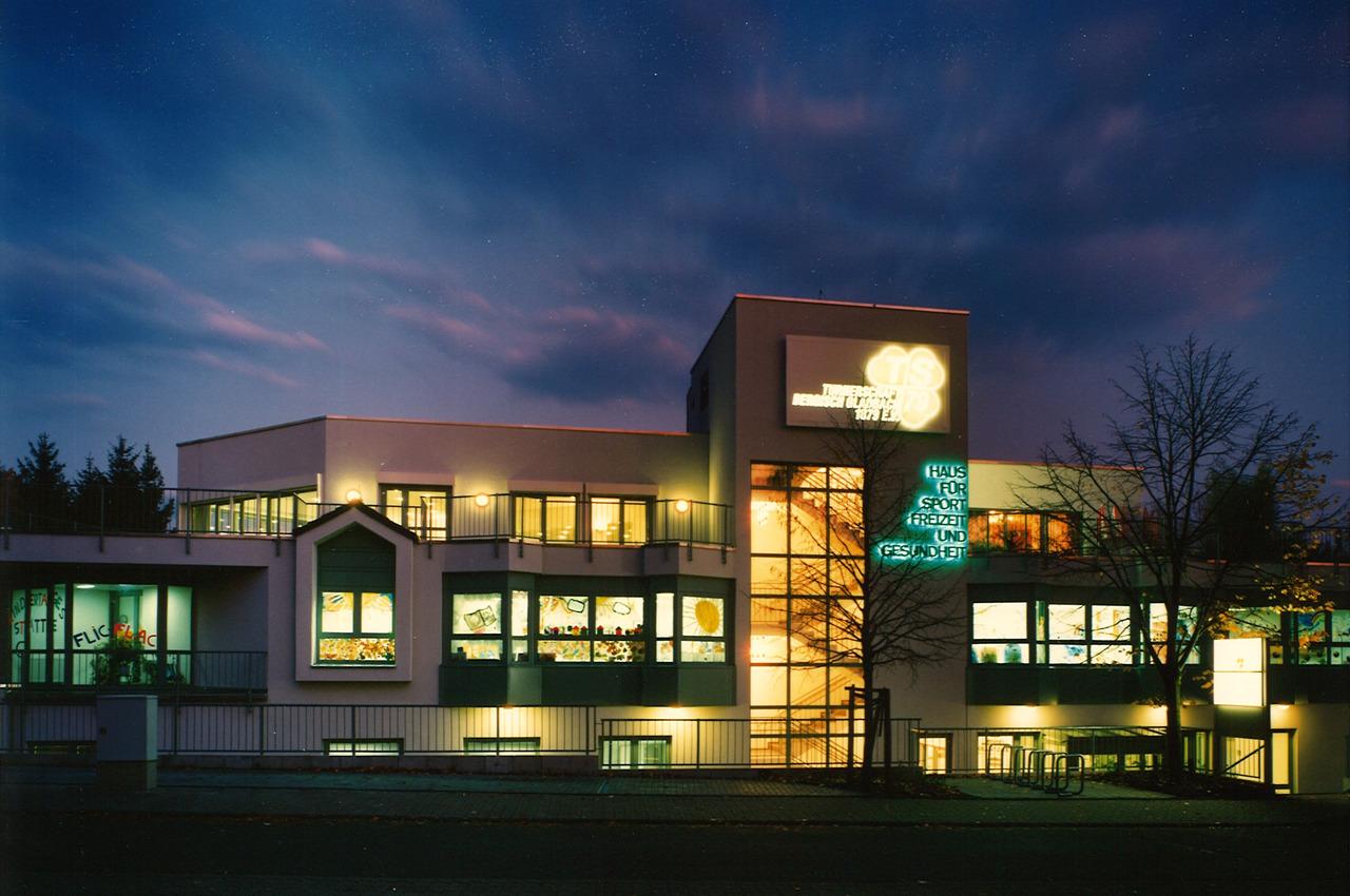 Moderne Sporteinrichtung bei D&auml;mmerung, mit beleuchteten Fenstern und dem Schriftzug &bdquo;Haus f&uuml;r Sport, Freizeit und Gesundheit&ldquo;.