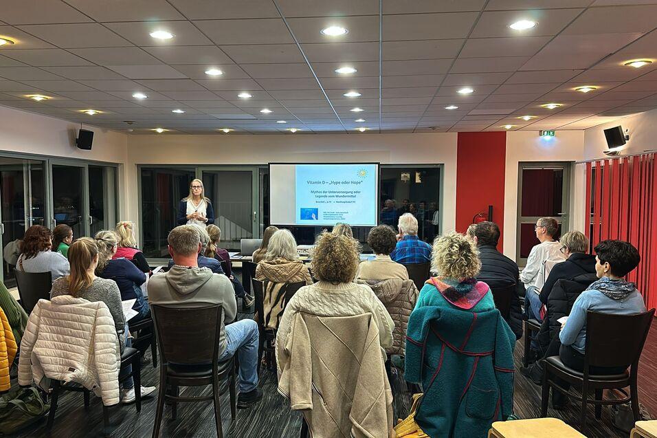 Eine Referentin steht vor einem Vortrag, während eine Gruppe von Personen sitzend aufmerksam zuhört.