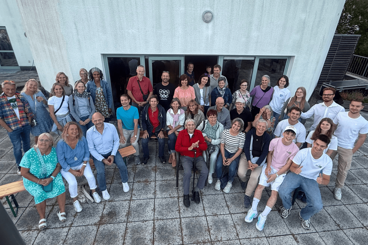Gruppe von etwa 30 Personen steht lächelnd vor einem hellen Gebäude auf einem Flachdach.