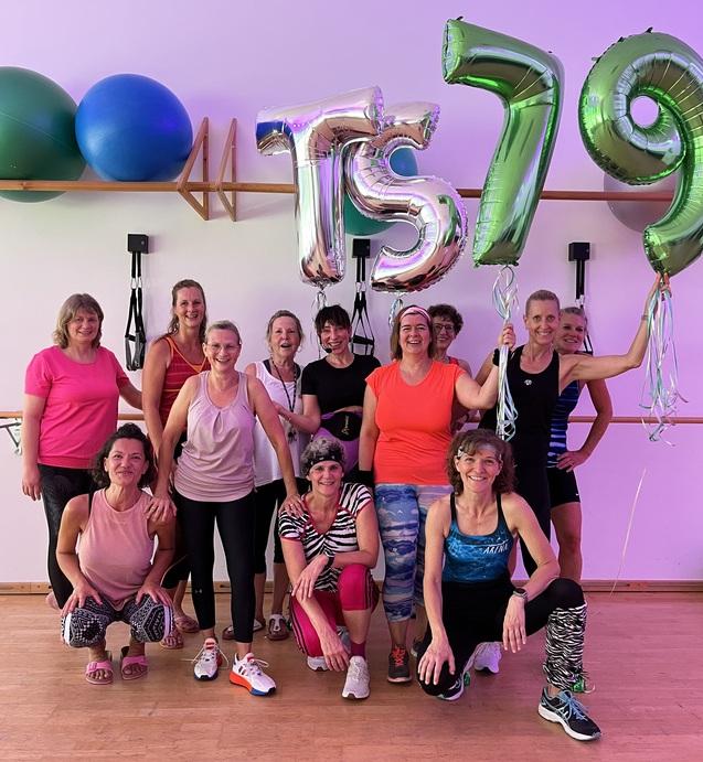 Gruppe von 12 Frauen in Sportkleidung posiert vor bunten Luftballons mit der Aufschrift „TS 79“ in einem Fitnessraum.