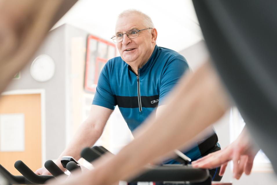 Älterer Mann mit Brille lächelt beim Radfahren in einem Fitnessraum, im Vordergrund Hände an Fahrradlenkern.