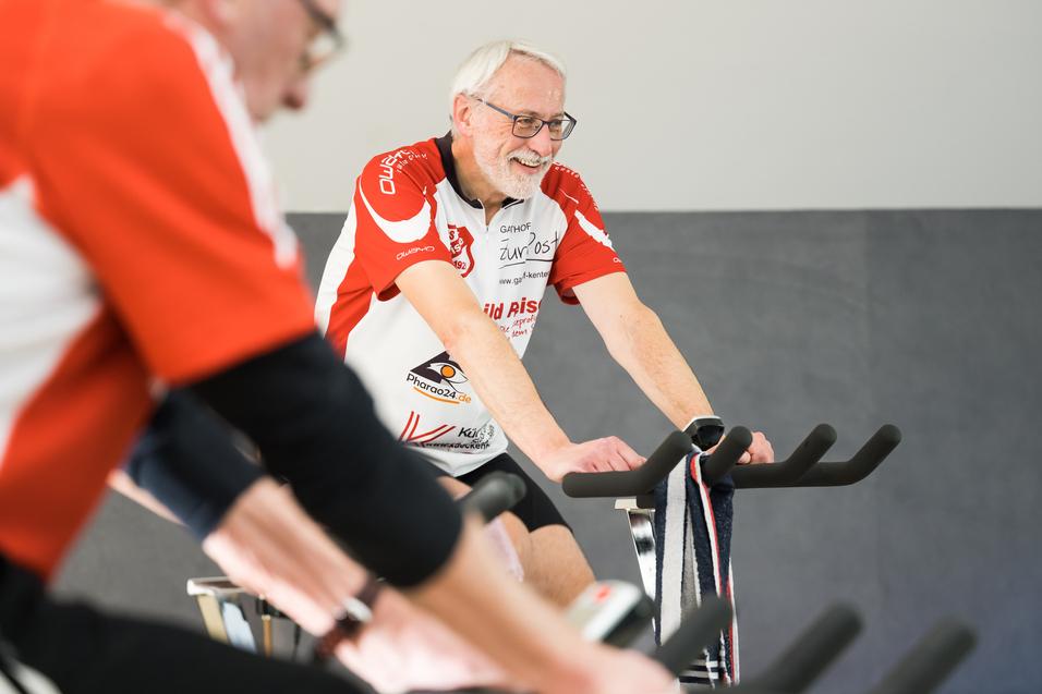 Zwei Radfahrer in Trainingskleidung lächeln auf stationären Fahrrädern während eines Fitnesskurses in einem Studio.