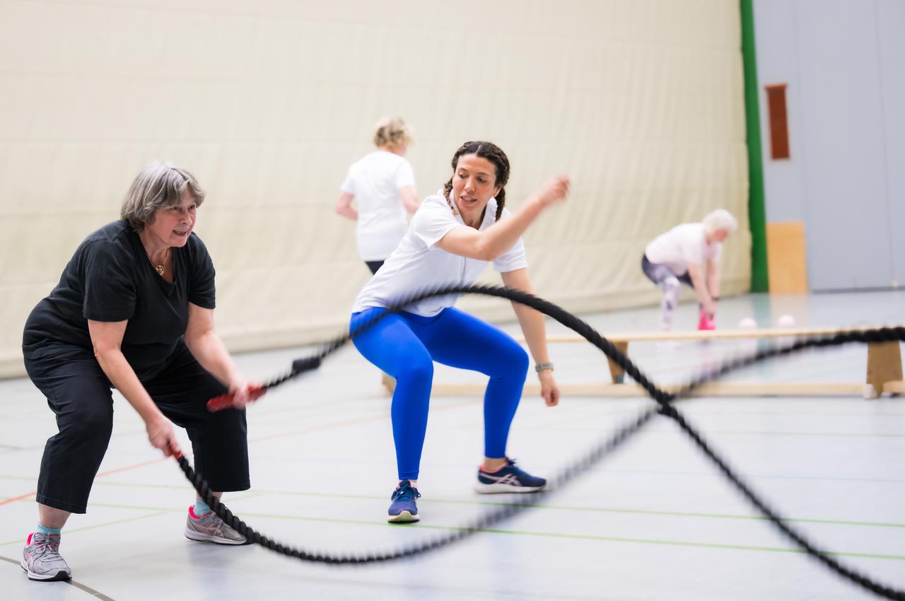 Zwei Frauen trainieren mit einem Battle Rope in einer Sporthalle, mehrere weitere Personen im Hintergrund aktiv.