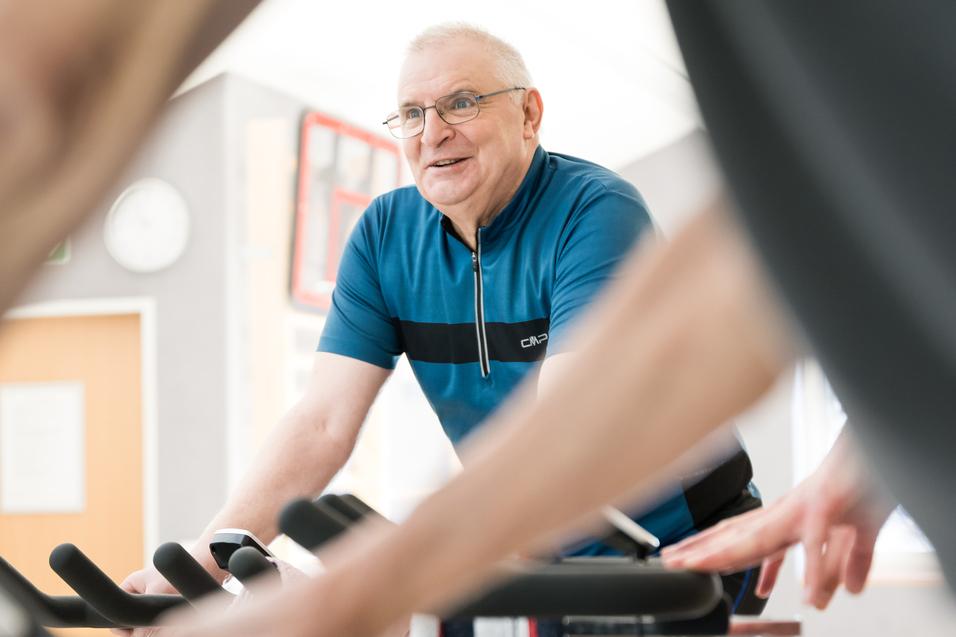 Älterer Mann mit Brille lächelt beim Radfahren in einem Fitnessraum, im Vordergrund Hände an Fahrradlenkern.