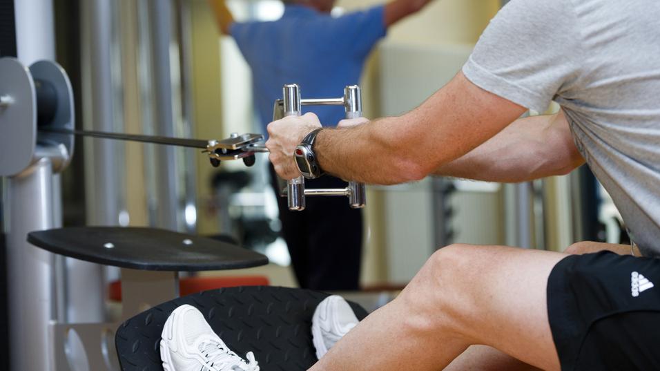 Mann trainiert im Fitnessstudio mit einem Kabelzuggerät, ein anderer Mensch im Hintergrund führt eine Übung aus.